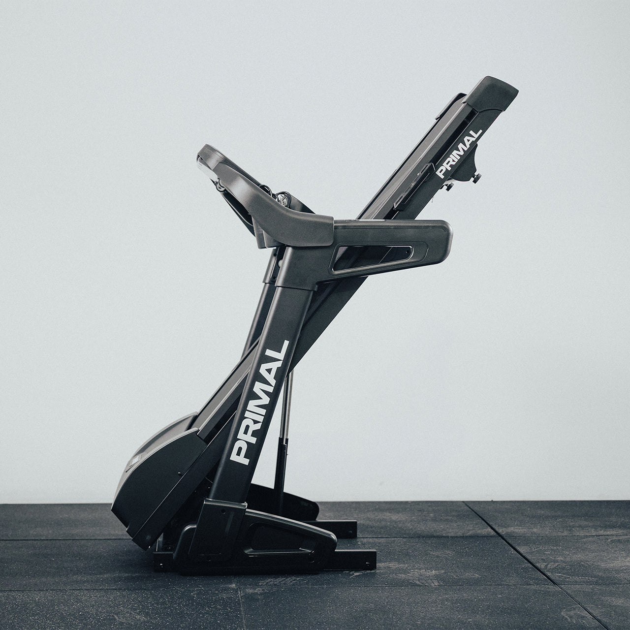 Black Primal folding treadmill on a grey floor with a white wall background