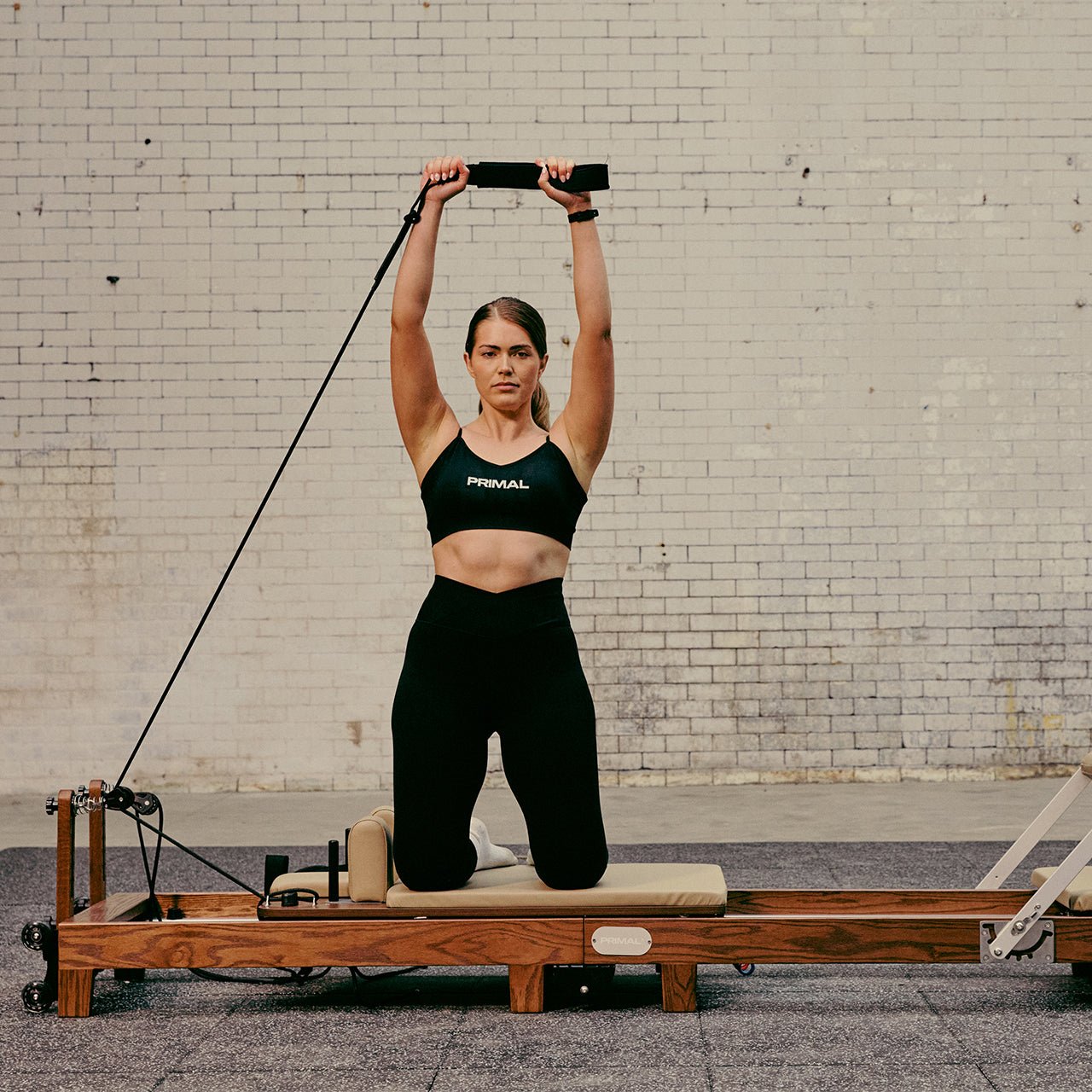 Woman exercising on a Pilates reformer machine against a brick wall.