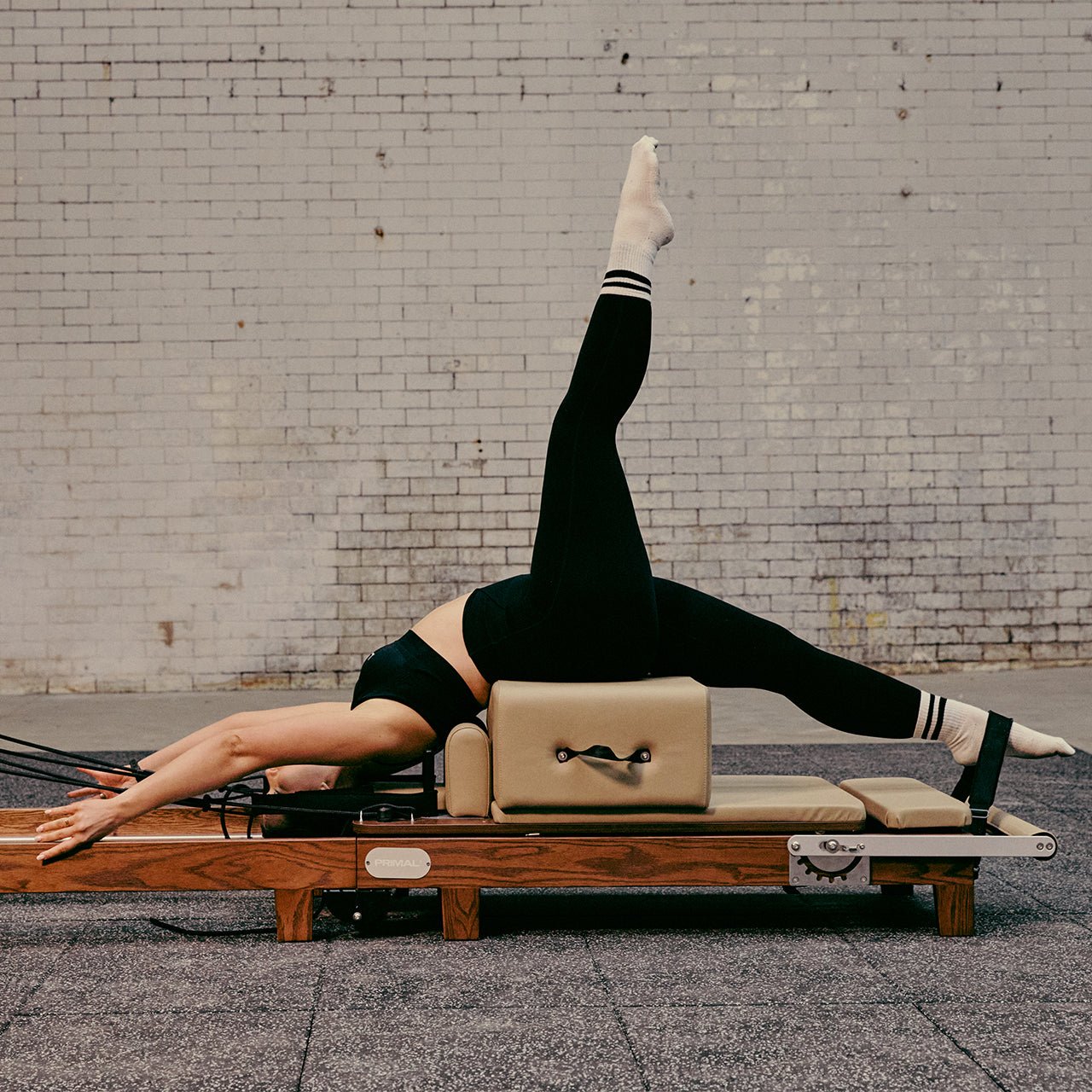 Person performing a Pilates exercise on a reformer machine against a brick wall.