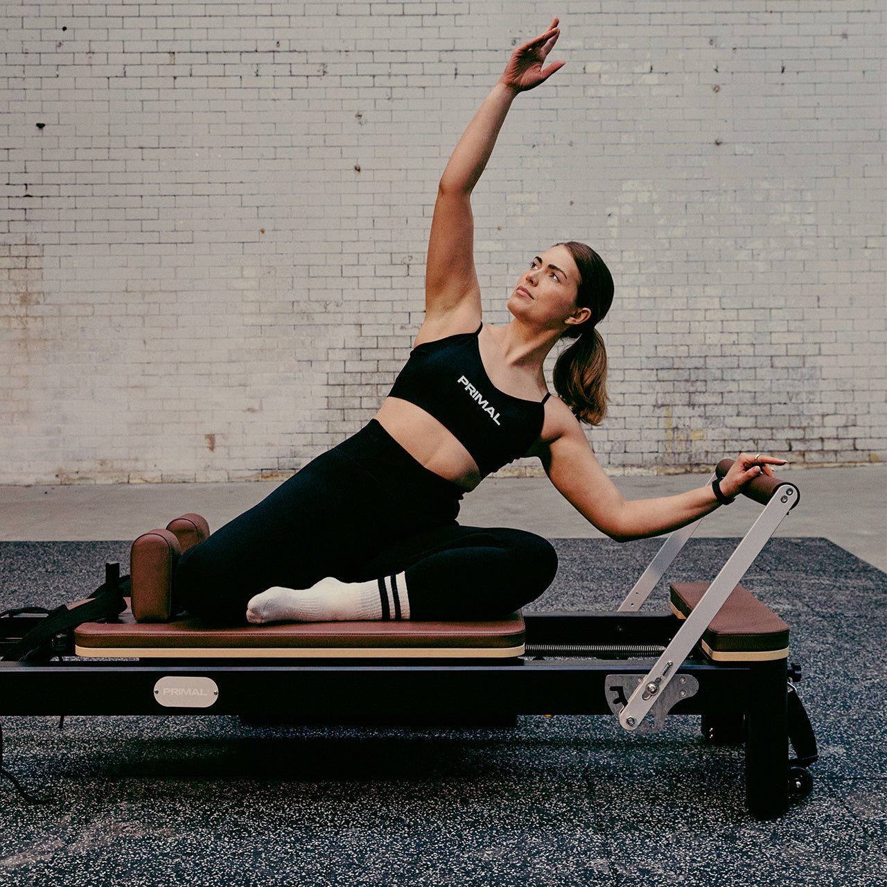 Woman exercising on a Pilates reformer machine against a brick wall.
